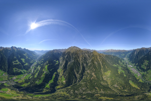 Panoramischer Blick auf ein Gebirgsmassiv von einem Hügel aus, mit grünem Gras, einer gewundenen Straße und einem hellblauen Himmel mit Sonnenlicht.