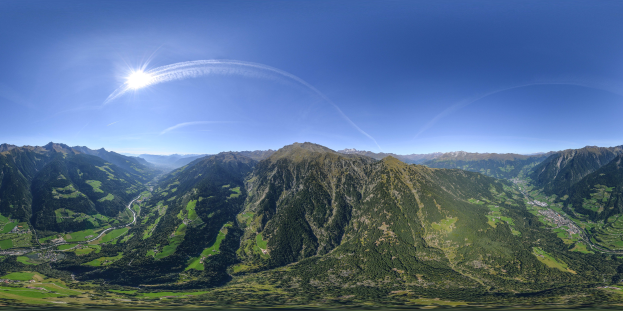 Panoramischer Blick auf ein Gebirgsmassiv von einem Hügel aus, mit grünem Gras, einer gewundenen Straße und einem hellblauen Himmel mit Sonnenlicht.