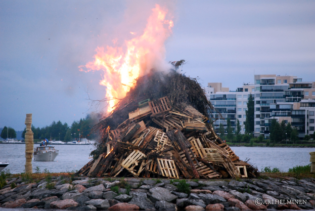 Holzobjekte brennen mit Feuer, mit Steinen am Boden, Wasser in der Mitte, einem Schiff auf dem Wasser und Gebäuden, Bäumen und Himmel im Hintergrund.