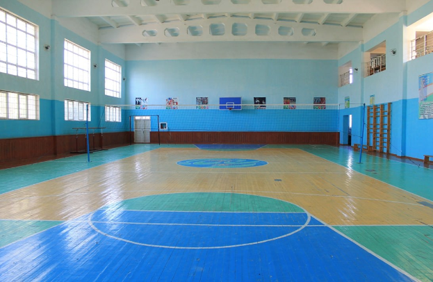 Großer Turnsaal mit blau-weißem Boden, einem Basketballfeld, einem Netz mit Pfosten, einer Tür, Fenstern, Wandplakaten, einem Geländer und einem Dach.