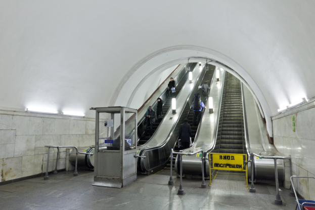 Rolltreppe in einer U-Bahn-Station mit Menschen, Geländern, Informationsschildern, Wandbeleuchtung und einem Einkaufswagen unten.