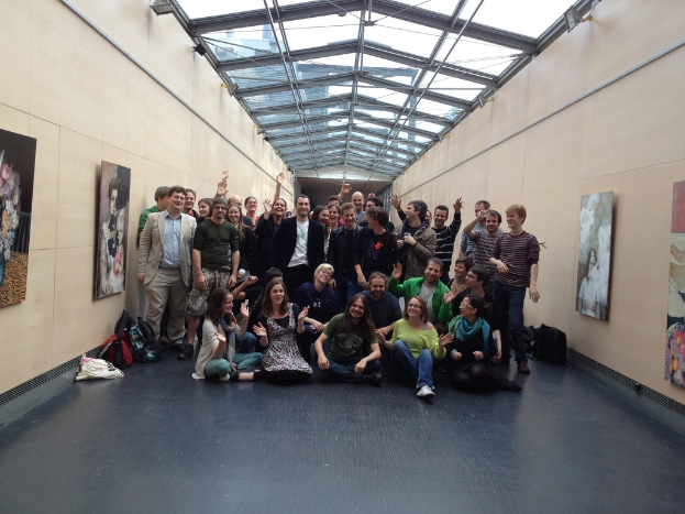 Eine Gruppe von Menschen, die für ein Foto posen, einige sitzen und einige stehen, mit Taschen auf dem Boden und gerahmten Gegenständen an den Wänden, unter dem Dach eines Unterschlupfs.