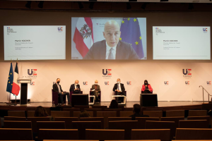 Eine Gruppe von Menschen sitzt auf einer Bühne in einem Halbkreis um ein Podium mit Mikrofon, gegenüber einem Publikum, mit Flaggen auf beiden Seiten und einem Bildschirm, der Text und Bilder anzeigt, im Hintergrund beleuchtet von Scheinwerfern bei einer Pressekonferenz der Europäischen Union.