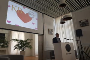 Ein Mann steht an einem Rednerpult mit einem Mikrofon, h├Ąlt eine Pr├Ąsentation über Geschlechtergerechtigkeit, vor einem Projektionsschirm und einem Lautsprecher mit St├Ąnder, mit Stühlen, einem Topf mit einer Pflanze, gerahmten Fotos, Vorh├Ąngen und Deckenleuchten im Hintergrund.