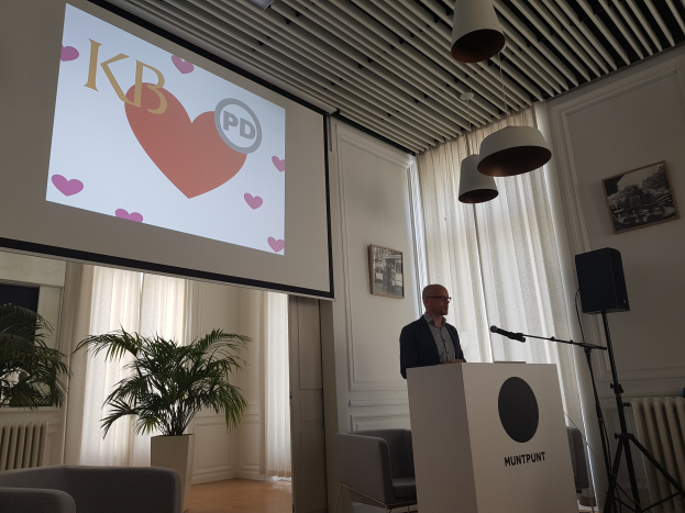 Ein Mann steht an einem Rednerpult mit einem Mikrofon, h├Ąlt eine Pr├Ąsentation über Geschlechtergerechtigkeit, vor einem Projektionsschirm und einem Lautsprecher mit St├Ąnder, mit Stühlen, einem Topf mit einer Pflanze, gerahmten Fotos, Vorh├Ąngen und Deckenleuchten im Hintergrund.