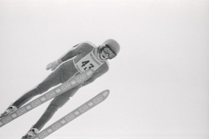 Ein Skispringer in voller Ausrüstung springt in der Luft auf Skiern vor einem klaren blauen Himmel.