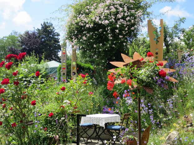 Ein Garten voller bunter Blumen und Pflanzen, mit einem Tisch mit einer Decke und Stühlen, vor Bäumen und einem bewölkten Himmel auf der Chelsea Flower Show.