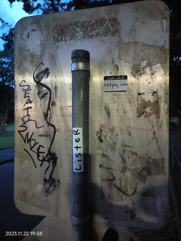 Ein Metallpfahl mit Graffiti steht in der Mitte einer Straße, umgeben von Bäumen, Gras und einer Straße, mit einem Himmel oben und einem Wasserzeichen links unten.