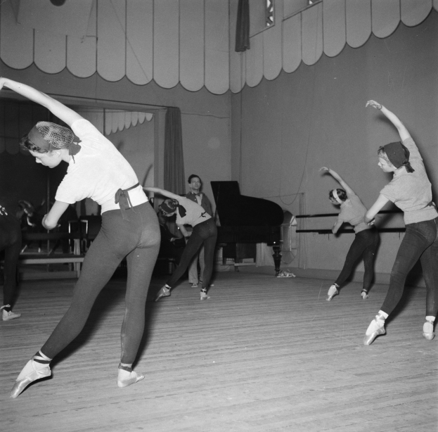 Eine Gruppe junger Frauen tanzt anmutig in einem schwarzen und weißen Tanzstudio, mit einem Klavier im Hintergrund und einer Wand dahinter.