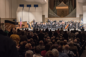 Eine große Gruppe von Menschen sitzt vor einer Bühne mit einigen, die Musikinstrumente halten. Es gibt Fahnen mit Stangen, einen Lautsprecherkasten und andere Gegenstände auf der Bühne, sowie eine Decke, Deckenleuchten und eine Tür an der Rückwand, was auf ein Konzert an der University of Texas at Austin hindeutet.