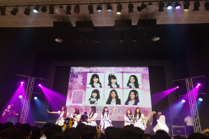 Eine Gruppe von Menschen performs auf einer hell erleuchteten Bühne mit Trägern, während eine Menge darunter das Ereignis aufzeichnet; ein großes Display im Hintergrund zeigt Bilder der K-Pop-Gruppe BK48.