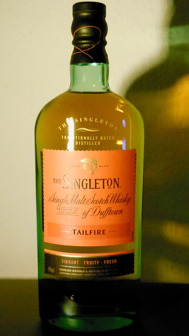 Eine tiefe bernsteinfarbene Flasche Singleton of Dufftown Tailfire Single Malt Scotch Whisky mit einem schwarzen Etikett, das ein Löwen- und Schlosswappen zeigt, auf einem Tisch.