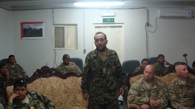 Eine Gruppe von Soldaten, die auf Stühlen und einer Couch sitzen, wobei einer steht und zu ihnen spricht.