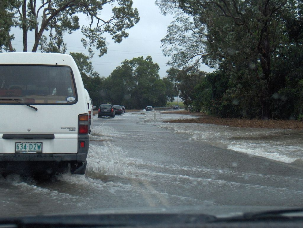 Eine Straße mit Wasser darauf, mehrere Autos, Bäume an den Seiten und ein bewölkter Himmel darüber.