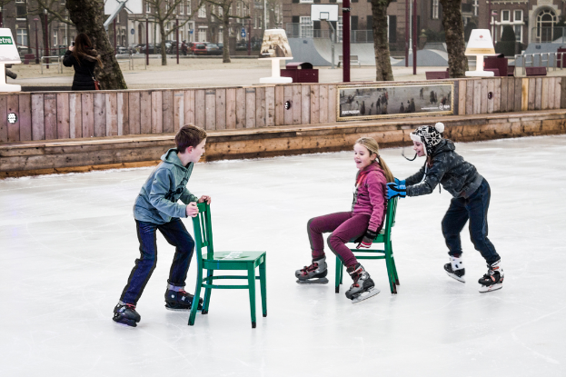 Kinder beim Skifahren im Vordergrund eines Spielplatzes mit drei Kindern und zwei Stühlen in der Mitte und Gebäuden, Bäumen, Bänken, Pfählen und einem Basketballfeld im Hintergrund.
