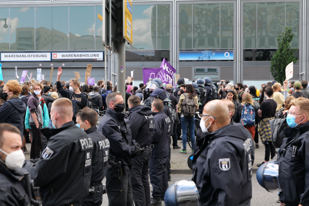 Große Menschenmenge vor einem Gebäude protestierend, einige mit Schildern und Helmen, mit einem Schildständer im Vordergrund und einem Baum im Hintergrund.