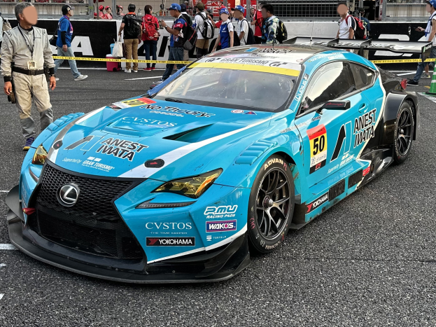 Blauer Lexus RC F GT3 Rennwagen auf einer Straße mit Zuschauern, einem Verkehrskegel rechts und Hintergrundwerbung und Geländern.