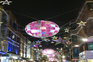 Eine nächtliche Stadtstraße mit Lichtern und einer großen Diskokugel, die von einem Gebäude hängt und eine festliche Atmosphäre schafft.