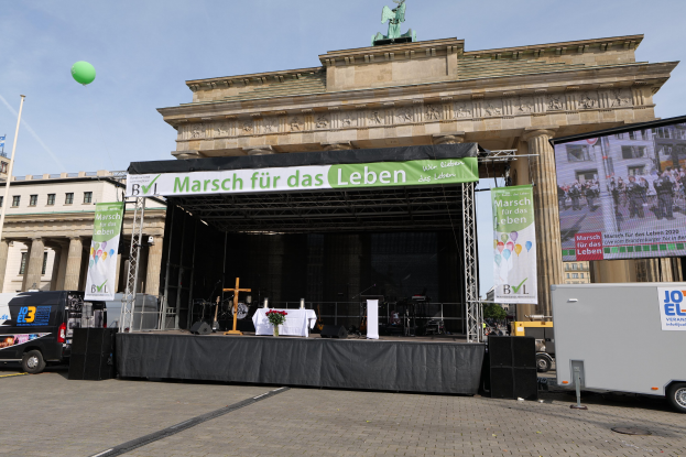 Eine Bühne vor dem Brandenburger Tor in Berlin, Deutschland, mit einem Tisch mit einer Blumenvase, einem Kreuzsymbol, Bannern mit Text, Rednern und anderen Gegenständen, sowie Fahrzeugen auf der Straße und Gebäuden im Hintergrund.
