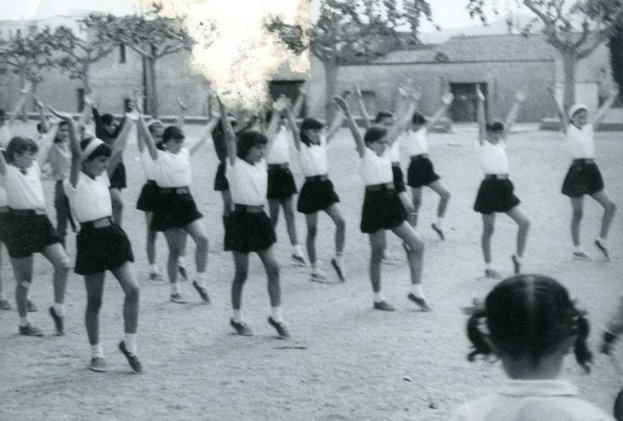Schwarzes und weißes Foto von jungen Mädchen in Schuluniformen, die auf einer Straße tanzen, die von Gebäuden und Bäumen gesäumt ist, mit einem Paar Schuhe am Boden.