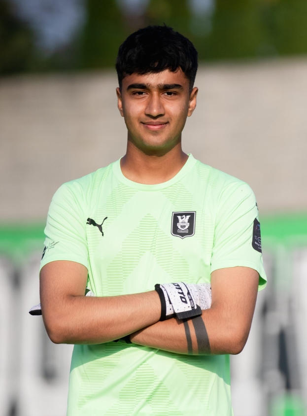 Ein junger Mann in einem grünen T-Shirt steht auf einem Fußballfeld und schaut mit verschränkten Armen und einem Lächeln in die Kamera, während der Hintergrund unscharf ist und darauf hindeutet, dass er im Fokus der Aufnahme steht.