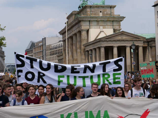 Gruppe von Studenten marschiert in Berlin mit einer leuchtend bunten "Students for Future"-Schlagzeile vor dem Hintergrund von Gebäuden, Bäumen und Himmel.