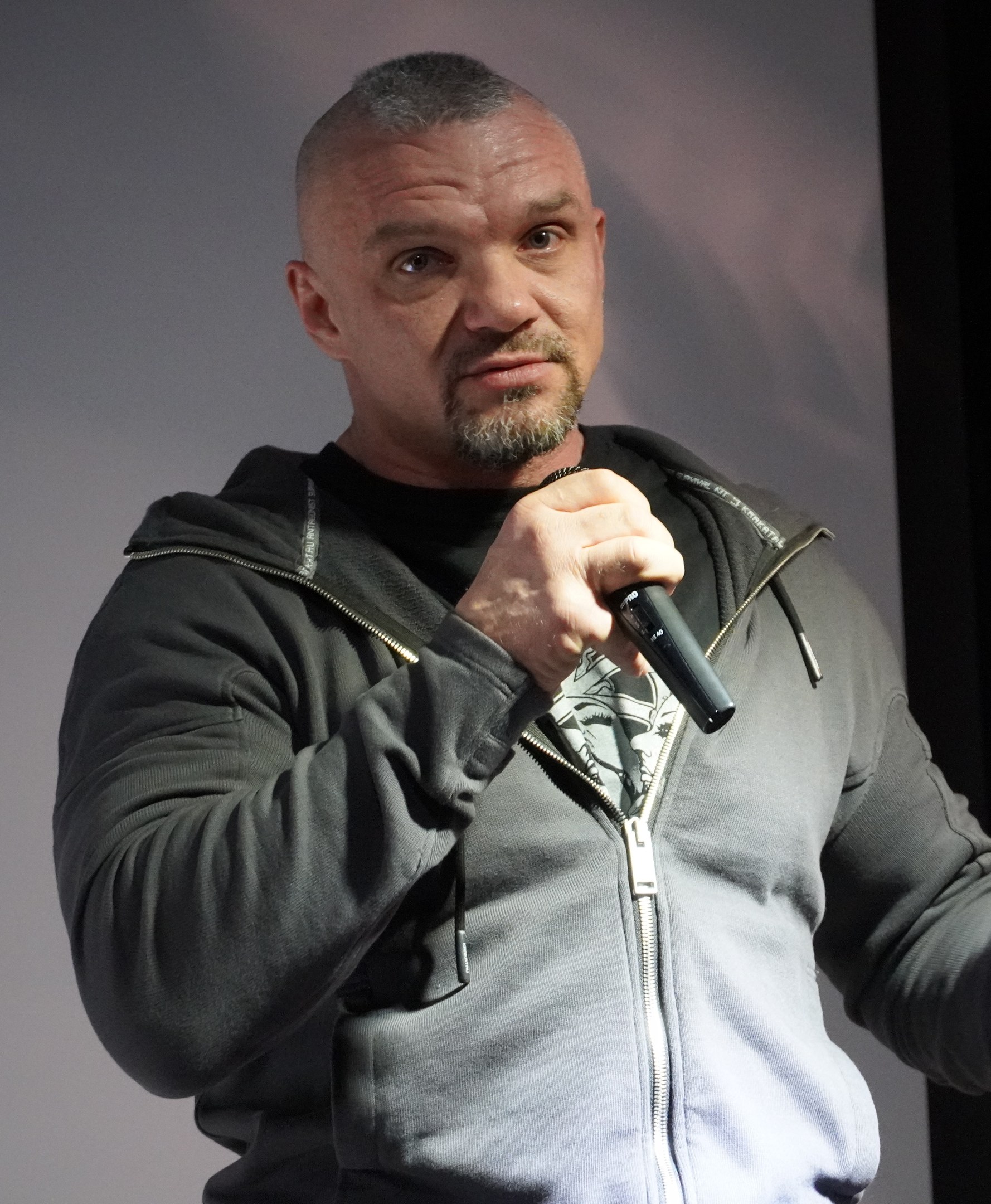 Ein Mann in einer schwarzen Kapuzenjacke und einem schwarzen Anzug mit einem weißen Hemd hält ein Mikrofon und steht selbstbewusst vor einer Wand, er scheint ein Komiker zu sein.