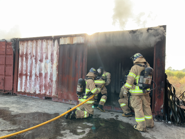 Feuerwehrleute in Schutzausrüstung löschen Feuer in einem Container, mit Rauchentwicklung und Grünzeug im Hintergrund.