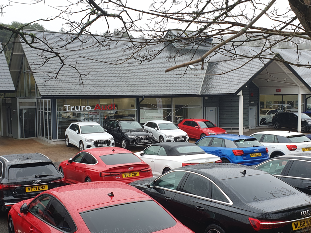 Ein Autohaus mit mehreren geparkten Autos, umgeben von Bäumen unter einem klaren blauen Himmel, mit einem Gebäude mit Dach, Glaswänden und einem Schild.