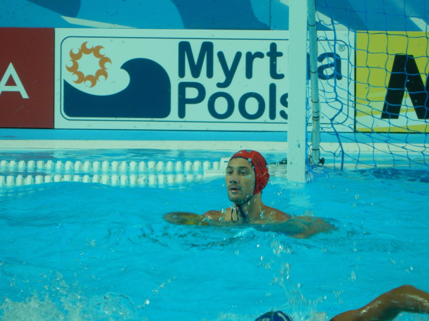 Wasserballspieler in Bewegung in einem Pool, trägt eine Mütze und hat ein Netz auf der rechten Seite und eine Werbetafel im Hintergrund.