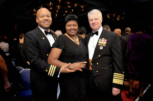 Zwei Männer in schwarzen Blazern und weißen Hemden stehen neben einer Frau in einem schwarzen Kleid, die eine Auszeichnung auf einem roten Teppich hält, während andere im Hintergrund unter beleuchteter Decke sitzen.