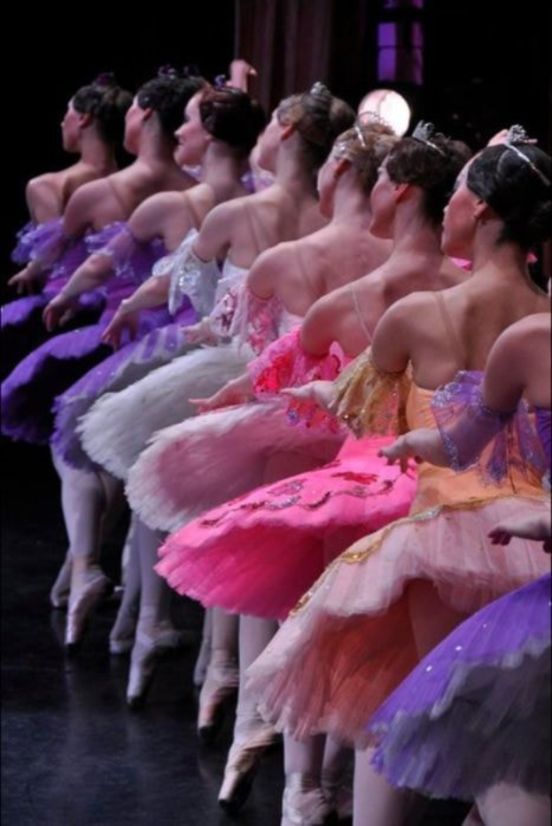 Balletttänzerinnen in rosafarbenen Tutus sitzen auf einer Bühne, beleuchtet von einem Licht im Hintergrund, bewegen sich synchron.