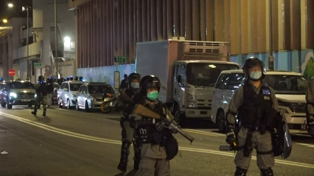 Polizeibeamte in Helmen, Handschuhen und Gesichtsmasken stehen nachts auf einer Straße, einige halten Waffen, mit Fahrzeugen, Laternenmasten, Schildern und Gebäuden im Hintergrund.