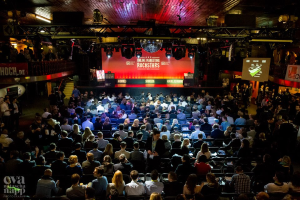 Eine große Menschenmenge sitzt auf Stühlen vor einer Bühne mit Bildschirmen und Bannern, beleuchtet von Scheinwerfern und umgeben von Geländern, voller Erwartung auf ein Online-Marketing-Rockstars-Event, mit einem Wasserzeichen in der linken unteren Ecke.