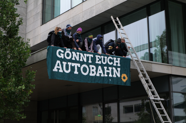 Gruppe von Menschen auf einem Gebäudedach mit einer Fahne, mit einem Baum links und Glasfenstern im Hintergrund, wahrscheinlich protestierend im Support der deutschen Regierung.