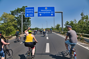 Gruppe von Radfahrern mit Helmen, die eine Straße mit einem Geländer und Bäumen auf beiden Seiten sowie Laternen im Hintergrund und einem klaren blauen Himmel entlangfahren, mit einem Schild oben, das eine Radtour in Hamburg anzeigt.