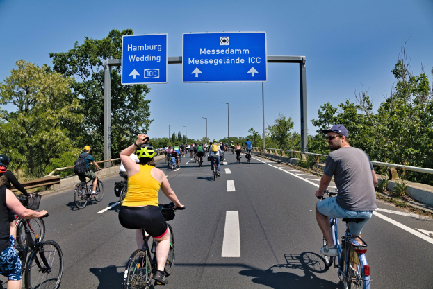 Gruppe von Radfahrern mit Helmen, die eine Straße mit einem Geländer und Bäumen auf beiden Seiten sowie Laternen im Hintergrund und einem klaren blauen Himmel entlangfahren, mit einem Schild oben, das eine Radtour in Hamburg anzeigt.