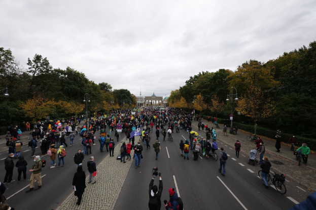 Eine große Gruppe von Menschen marschiert auf einer baumbestandenen Straße in Berlin, mit Kameras in der Hand, vor einem Gebäude und einem klaren Himmel.