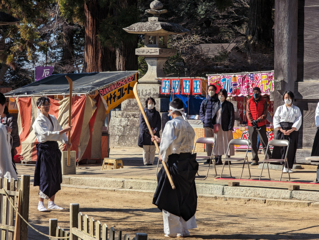 Eine Gruppe von Menschen in traditioneller Kleidung versammelt sich im Freien in Kyoto, einige tragen Masken und halten Holzstöcke, mit Stühlen, Bannern, einem Zelt und Bäumen im Hintergrund vor einem klaren blauen Himmel.