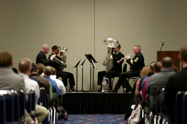 Eine Gruppe von Männern, die auf einer Bühne Blasinstrumente spielen, mit Notenständern, einem Podium mit Mikrofon und einem Publikum, das auf Stühlen sitzt, mit einer Wand im Hintergrund.