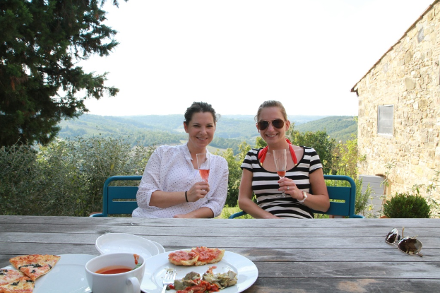 Zwei Frauen sitzen auf Stühlen im Freien, lächeln und halten Gläser in den Händen, mit einem Tisch zwischen ihnen, auf dem Goggles, ein Becher und ein Teller mit Pizza-Scheiben mit einer Gabel zu sehen sind.