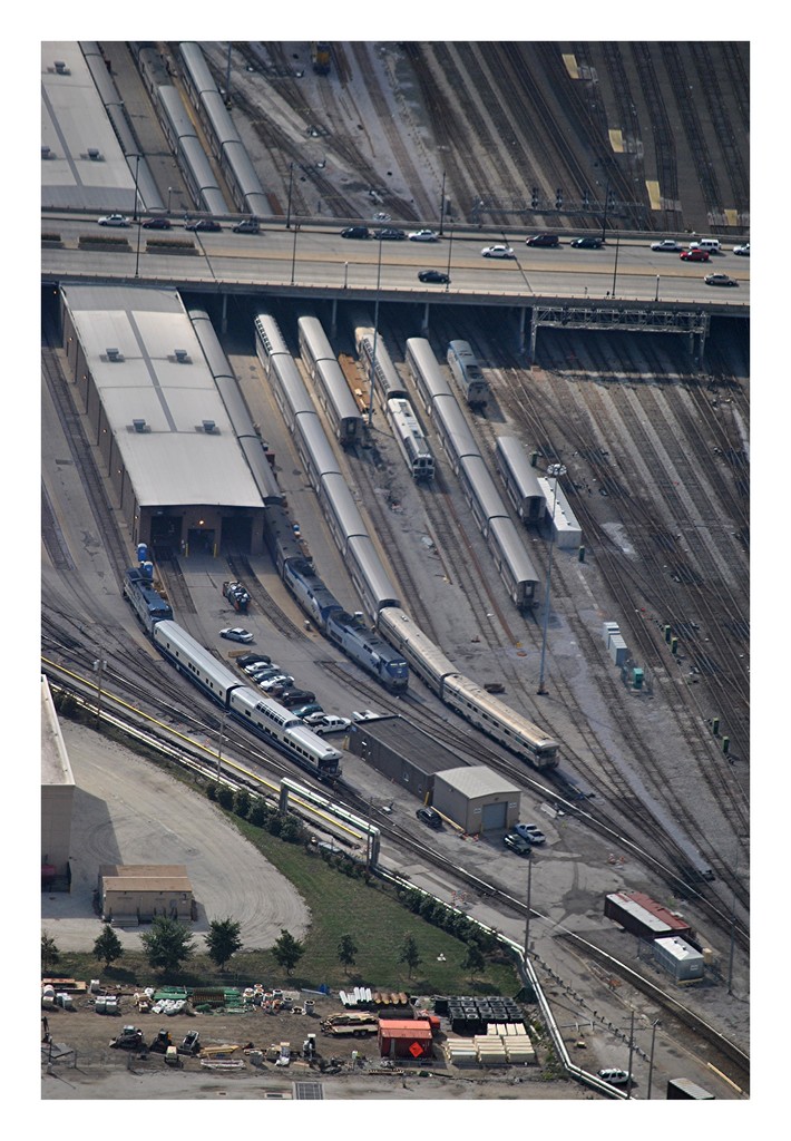 Eine Brücke mit wenigen Fahrzeugen darauf, Eisenbahnschienen mit wenigen Zügen darunter und eine Landschaft mit Gebäuden, Bäumen, Masten und diversen Gegenständen und Fahrzeugen am Boden.
