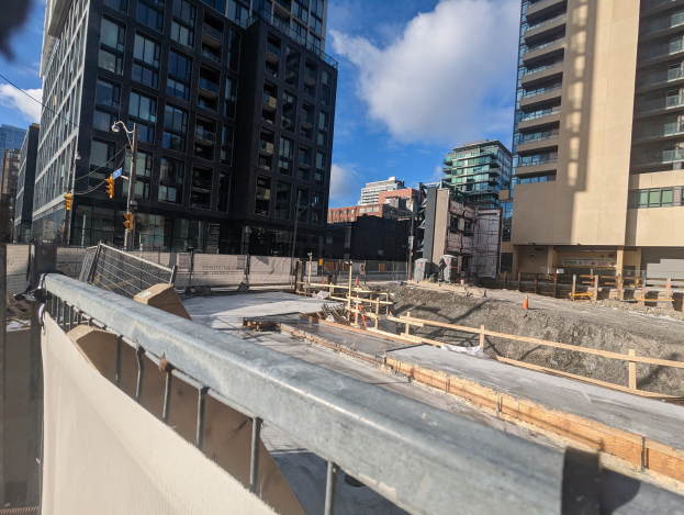 Baustelle in Toronto, Ontario, mit einem großen Gebäude im Hintergrund, umgeben von Sicherheitsbarrieren, Pfählen, Verkehrszeichen und Schildern unter einem bewölkten Himmel.