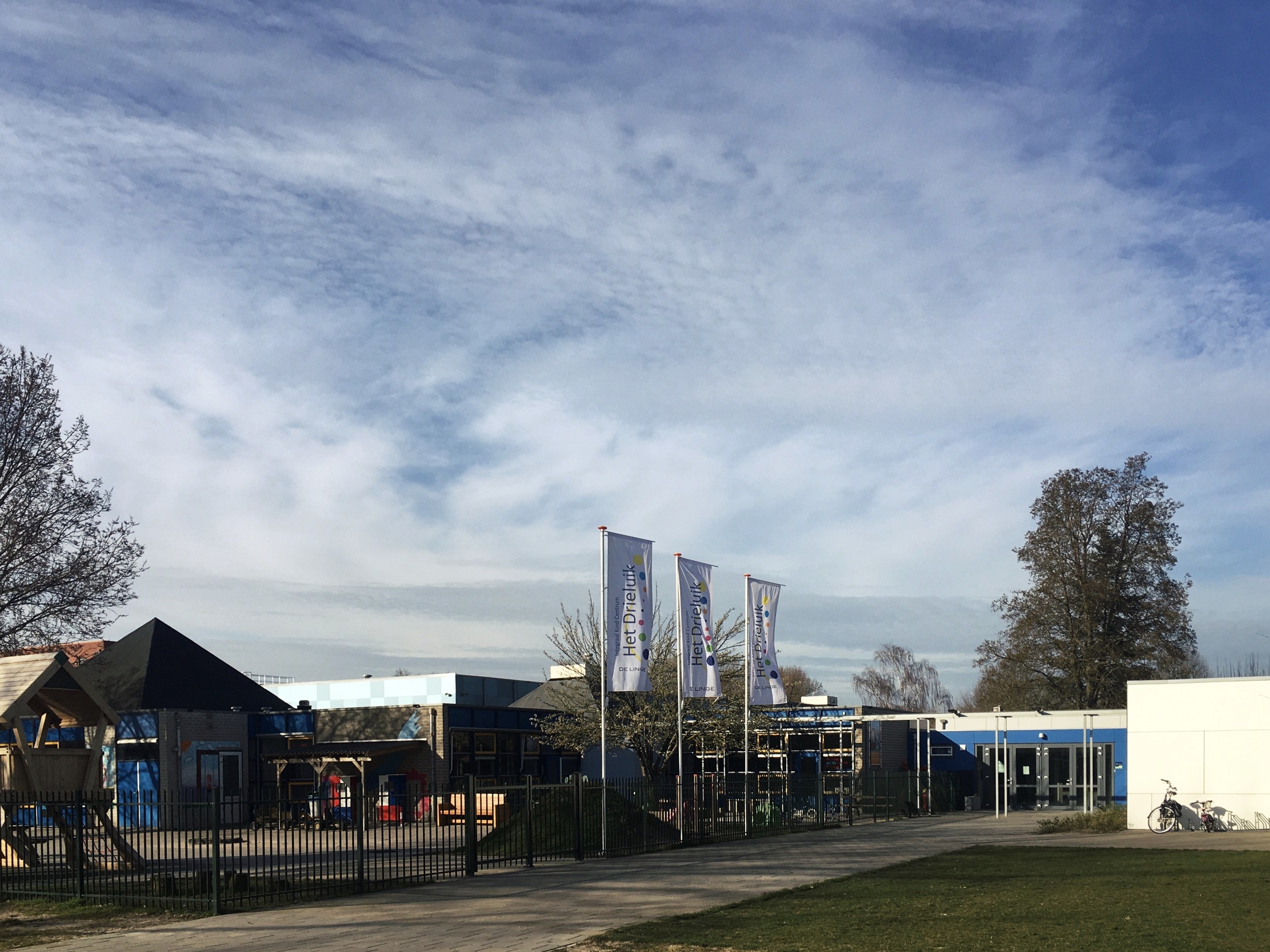 Großes Gebäude mit Metallzaun, Rasenfläche, Weg, Bäumen, Pfählen, Bannern, einem abgestellten Fahrrad, nahen Häusern und einem bewölkten Himmel, identifiziert als der neue Spielbereich für Kinder an der Schule.