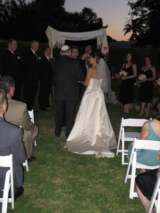 Eine Gruppe von Menschen steht bei einer Hochzeit, wobei Frauen rechts Blumensträuße halten, während andere auf Stühlen sitzen und Bäume im Hintergrund zu sehen sind.