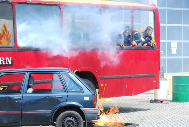Roter Doppeldeckerbus mit Rauch, daneben geparktes Auto mit drei sichtbaren Insassen und einem Gebäude mit Glasfenstern und einem Fass auf der rechten Seite im Hintergrund.