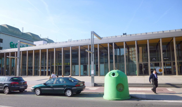 Grüner Müllcontainer auf einer Stadtstraße neben einem gläsernen Gebäude mit Säulen, mit Fahrzeugen und Fußgängern im Hintergrund unter einem bewölkten Himmel.