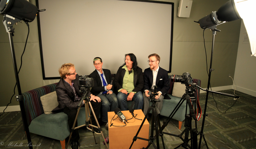Eine Gruppe von drei Personen sitzt auf einem Sofa und gibt ein Interview. Links sitzt eine Person in schwarzer Jacke und Hose, in der Mitte eine Person in Jacke und blauer Krawatte mit einem Mikrofon und rechts eine strahlende Person. Auf dem Tisch in der Mitte steht ein Mikrofonrecorder, Kameras sind links und rechts zu sehen und große Scheinwerfer sind vorhanden. Im Hintergrund ist ein Projektionsschirm zu sehen.