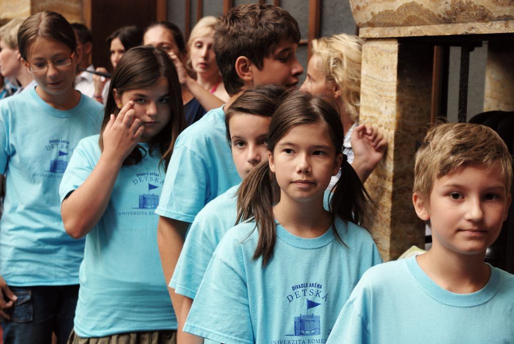 Eine Gruppe von Kindern steht vor einer Wand oder einer Säule, mit weiteren Personen dahinter und zusätzlichen Säulen und Fenstern im Hintergrund.