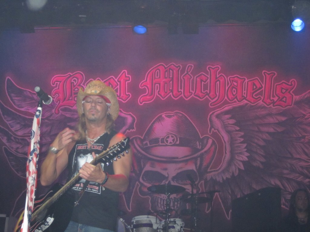 Eine Musikkonzertszene mit einem Gitarristen auf der Bühne, einer Person in der unteren rechten Ecke, einem Banner im Hintergrund und Drogen in der Mitte unten, mit Bühnenbeleuchtung von oben.